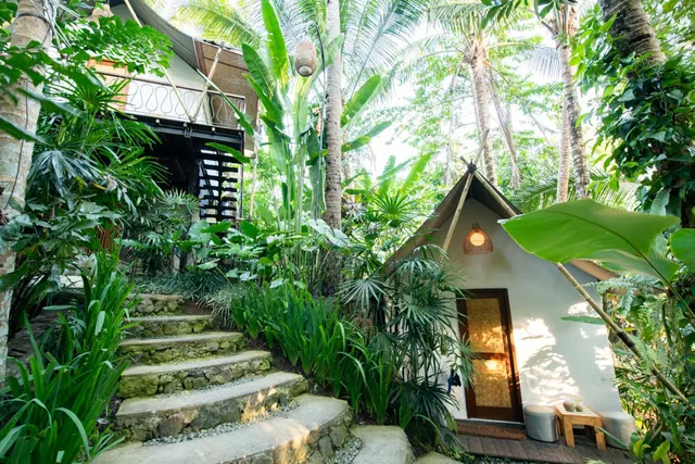 A small white cabin with a canvas roof and wooden door stands among palm trees and tropical plants, reached by a stone staircase.