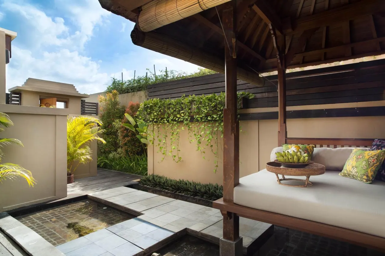A covered wooden daybed with cushions and a fruit bowl sits beside a small stone-edged water feature and a green plant-covered wall on a private outdoor terrace.