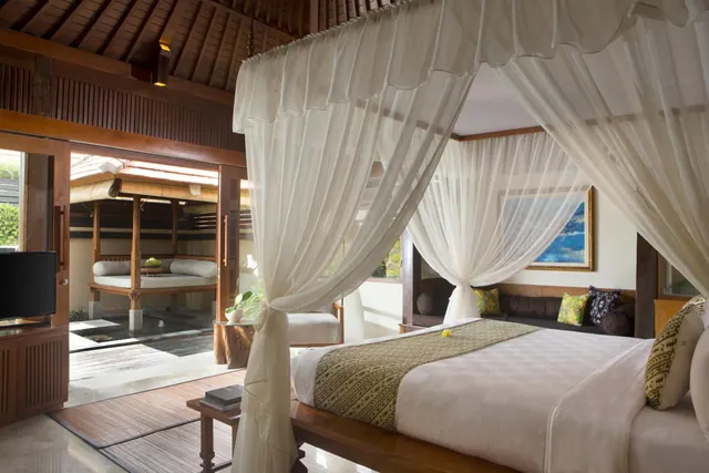 A bedroom with a canopy bed draped in sheer white netting, a cushioned wooden bench, a flat-screen television, and an open doorway to an outdoor pavilion with a daybed.