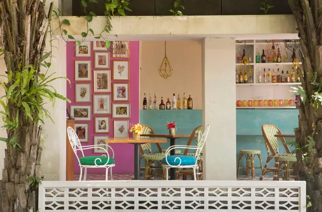 A bar area with pink wall displaying framed artwork, shelves of bottles and glasses, and bistro tables and chairs with coloured cushions, framed by palm trunks.