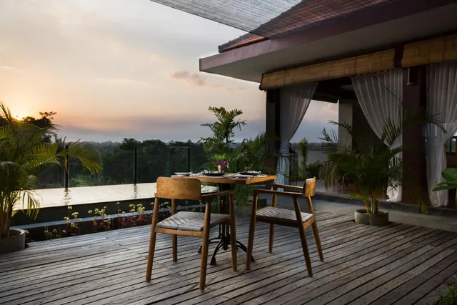 A wooden deck terrace with a small table and two chairs overlooks an infinity pool and tree-covered landscape at sunset.