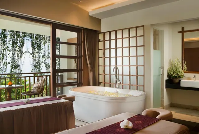 A spa treatment room with two massage tables, a freestanding bathtub with floating petals, a wooden lattice screen, and a balcony looking out onto greenery.