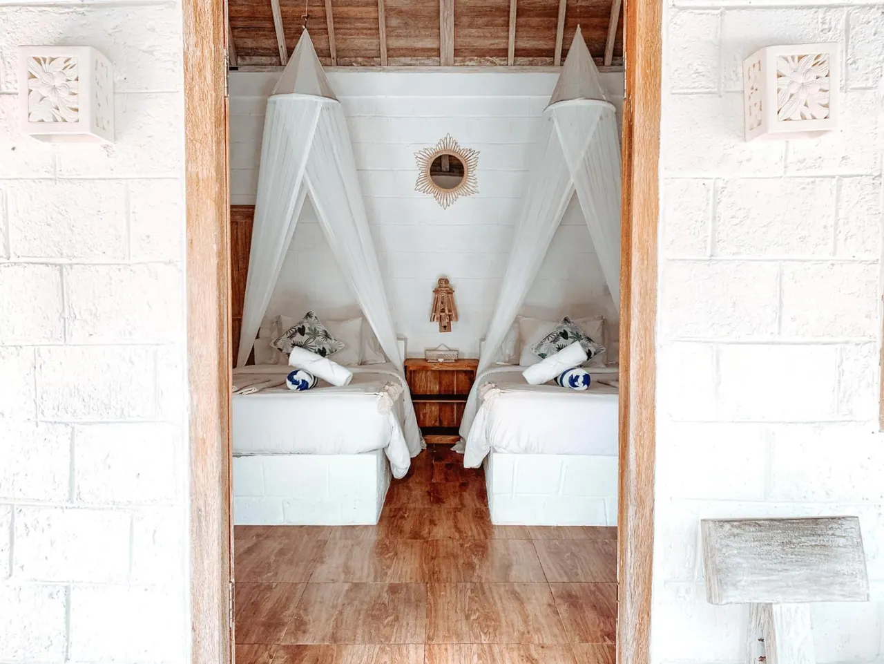 Two single beds with white mosquito nets and rolled towels stand side by side in a white-walled room with a timber floor, seen through a doorway.