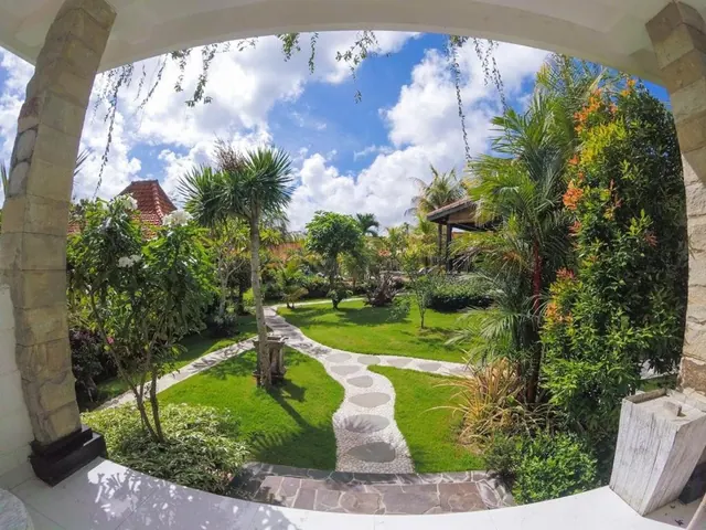 View through a stone archway from a covered terrace over a lawn with a stepping-stone path, palm trees and flowering shrubs under a partly cloudy sky.