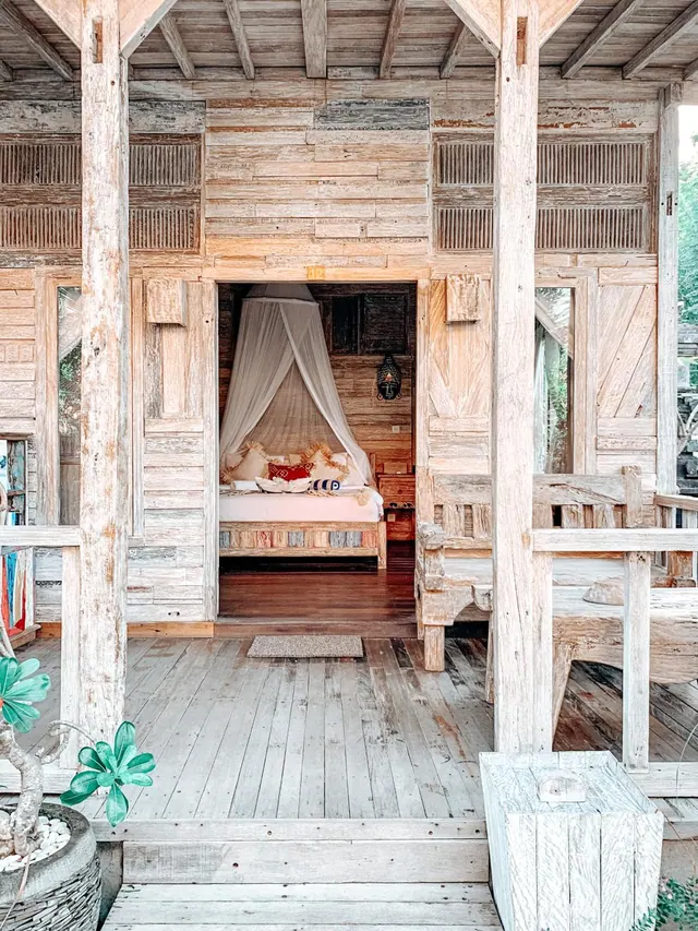 A wooden veranda with a carved facade leads through a doorway to a bedroom with a canopy bed under a mosquito net.