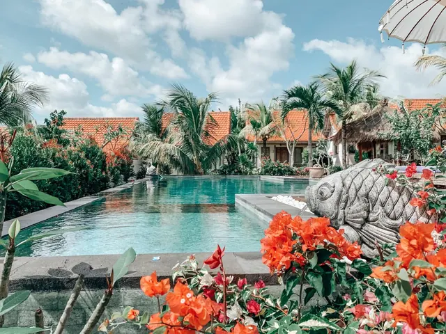 A long swimming pool with stone edging and a stone fish sculpture beside it, with red flowers in the foreground and orange-roofed villas and palm trees behind.