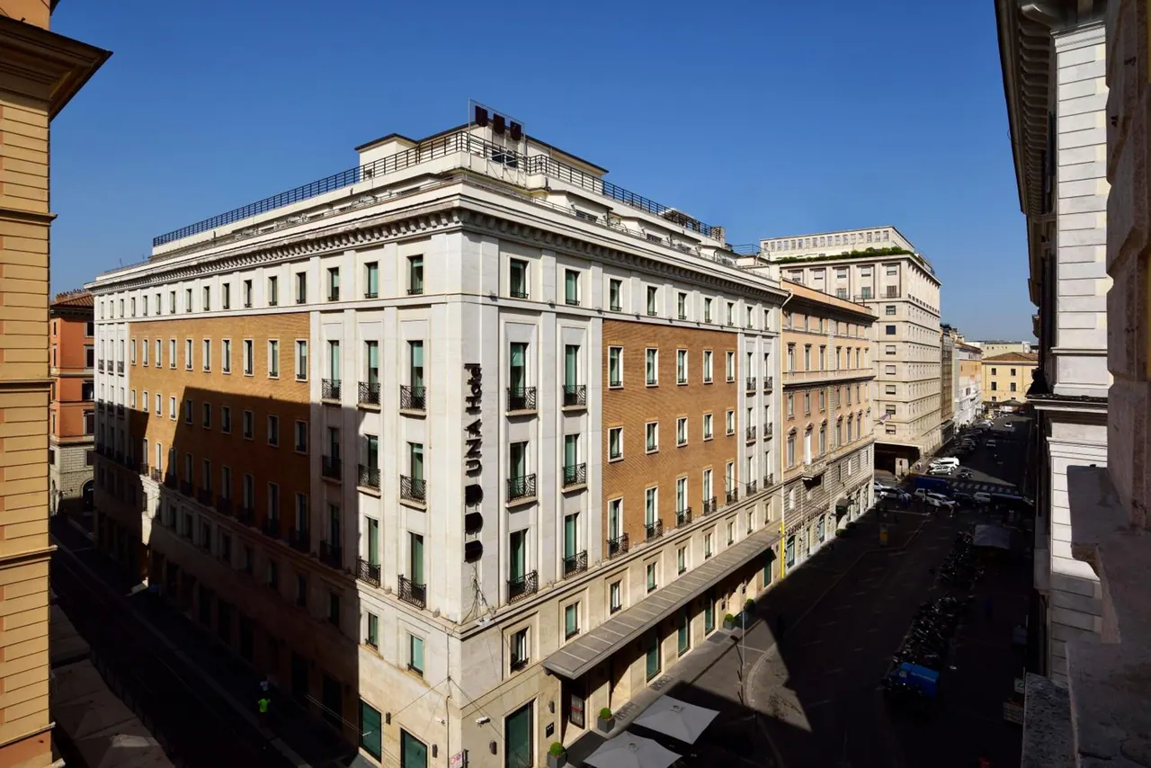 A cream and ochre multi-storey hotel building on a street corner in Rome, photographed from above under a clear blue sky.