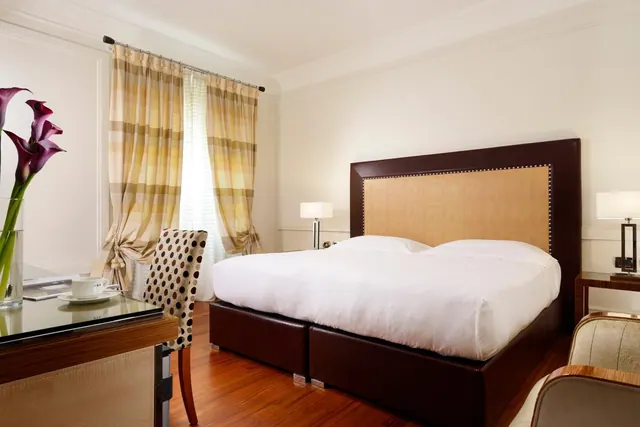 A bedroom with a large double bed with a dark upholstered headboard, bedside lamps, a glass-top desk with a cup and saucer, and sheer and striped curtains over a window.