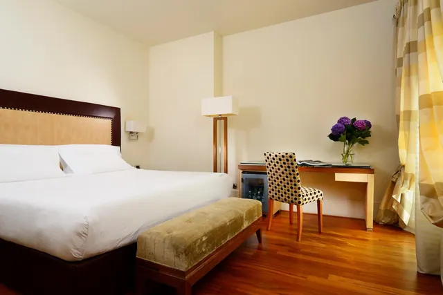 A bedroom with a large white bed, an upholstered bench at its foot, a wooden desk with a patterned chair and a small fridge beneath it, and a vase of purple flowers on the desk.