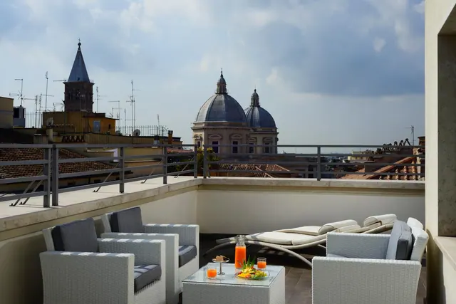 A rooftop terrace with wicker armchairs, a glass table holding juice and a fruit plate, sun loungers, and a view of church domes and a bell tower over the rooftops.