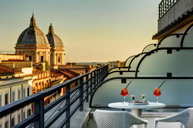 A rooftop terrace with a round table holding two cocktails and a small snack plate, overlooking domed churches and rooftops at sunset.