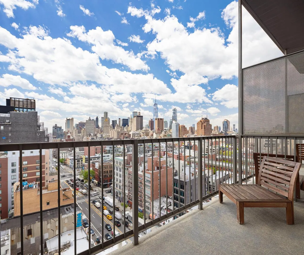 A private balcony with a wooden bench and metal railing overlooks a city street and skyline under a partly cloudy sky.