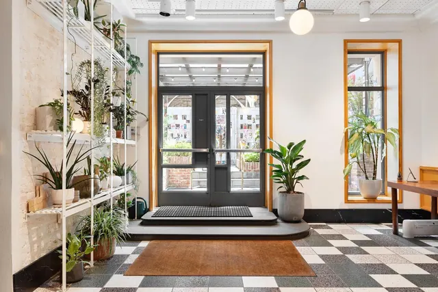 A lobby with a black and white checkered tile floor, potted plants on white metal shelving, a glass entrance door with a doormat, and a wooden table by a window.