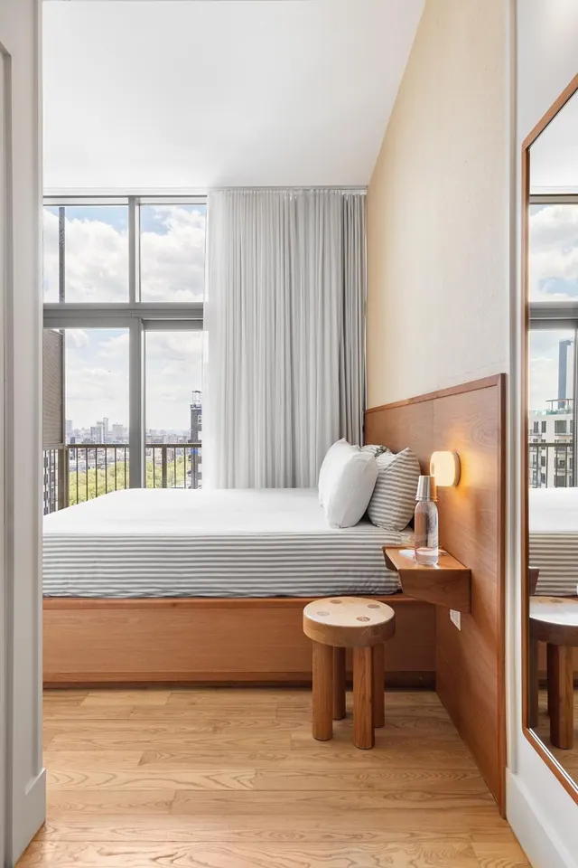 A bedroom with a made bed with grey striped linen, a wooden headboard and bedside shelf, and floor-to-ceiling windows looking out over trees and city buildings.