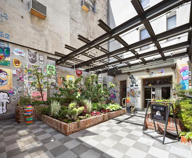A courtyard with a checkered tile floor, brick planter beds with shrubs, graffiti-covered walls, and a glass canopy overhead, with a welcome sign near the entrance.