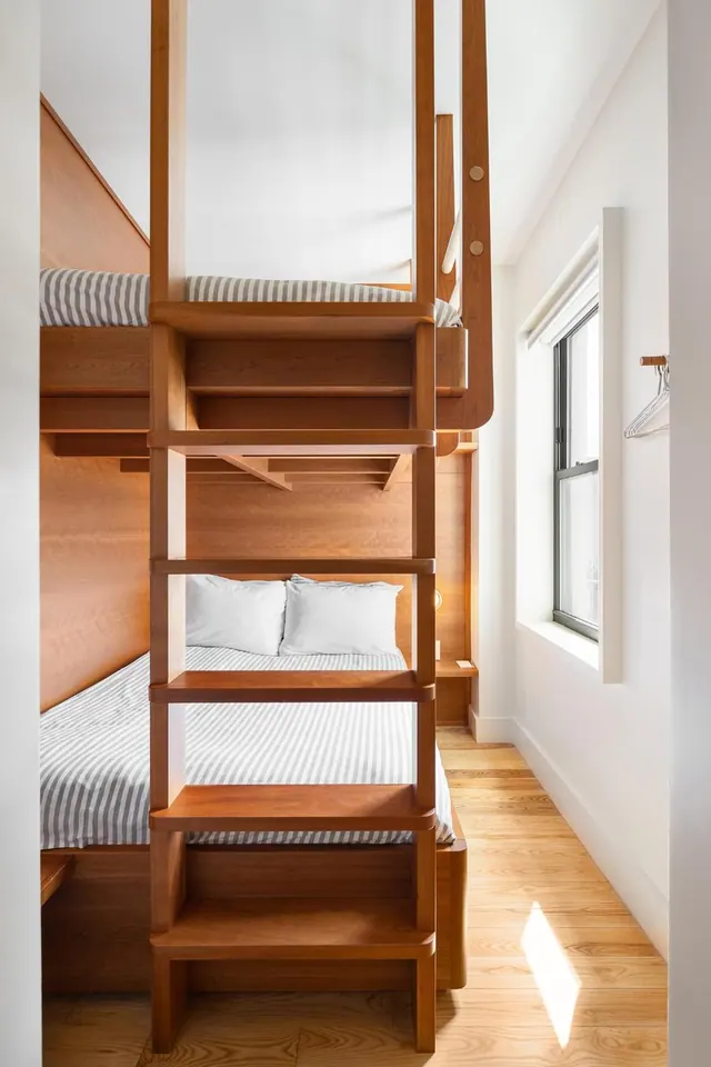 A small bedroom with built-in wooden bunk beds, a wooden ladder with shelves, grey striped bedding, and a window on the right wall.