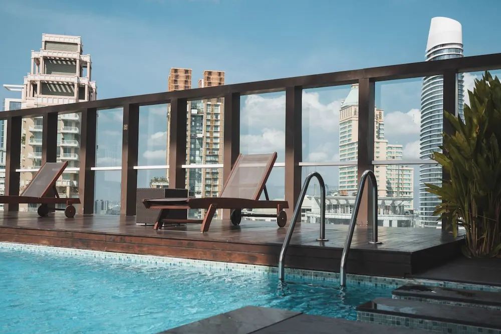 A rooftop swimming pool with wooden sun loungers beside a glass railing, overlooking high-rise towers under a blue sky.