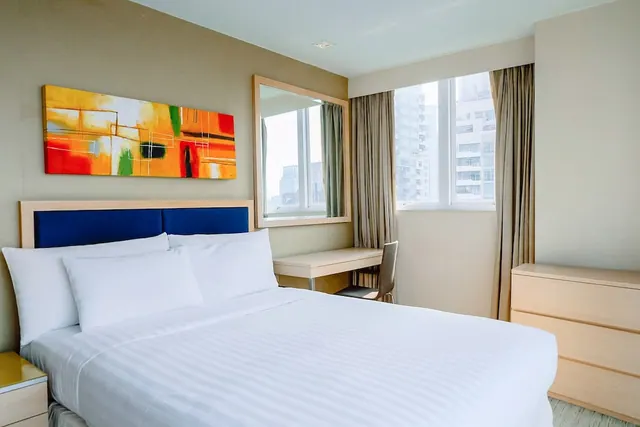 A bedroom with a king bed with a blue headboard, a colourful abstract painting, a mirror, a desk by the window and a view of high-rise buildings.