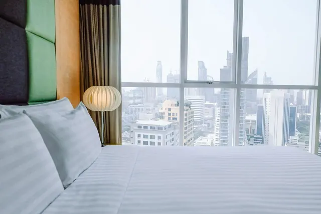 A made-up bed with striped white linen sits beside a lit bedside lamp, with floor-to-ceiling windows showing a hazy city skyline outside.