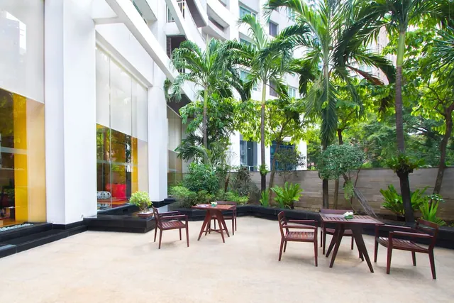 An outdoor paved courtyard with wooden tables and chairs, surrounded by palm trees and a white building with glass panels.