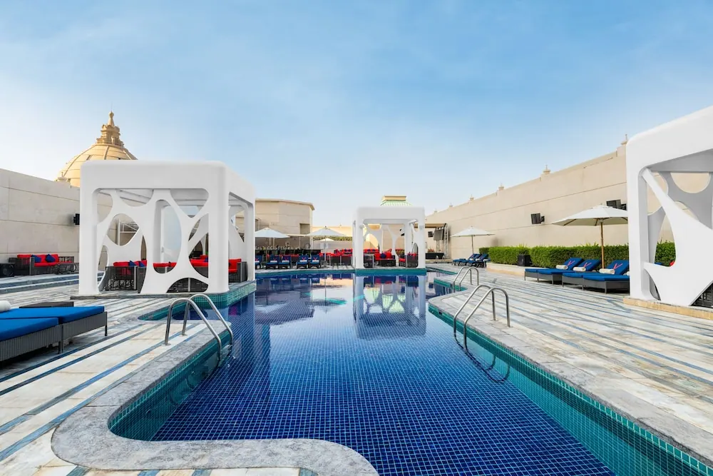A rooftop swimming pool with blue mosaic tiles, white cabanas, blue sun loungers and parasol umbrellas under a clear sky.