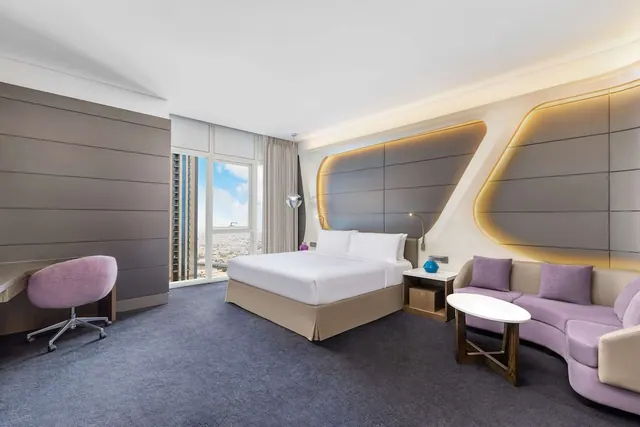 A hotel bedroom with a king bed, a purple sofa, a work desk, and floor-to-ceiling windows looking out over a city.
