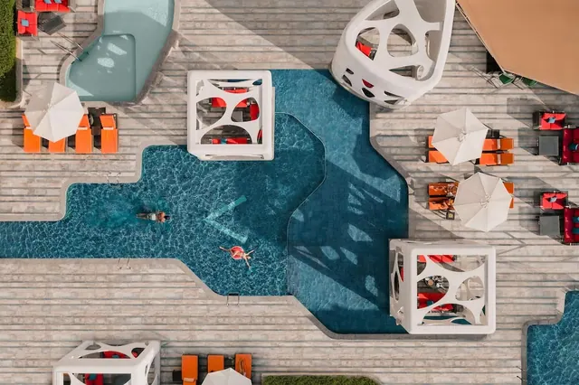 Aerial view of an irregularly shaped swimming pool with white sculptural cabanas, orange loungers, white umbrellas and two swimmers.