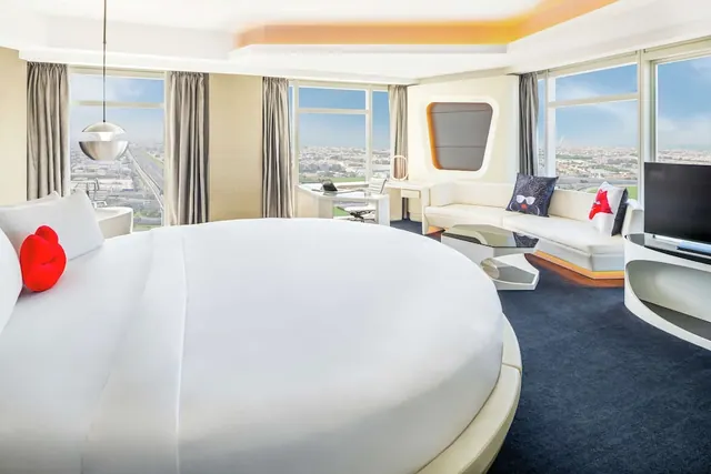 A bedroom with a large round bed, a white sofa, a desk and a television, with floor-to-ceiling windows looking over a city.