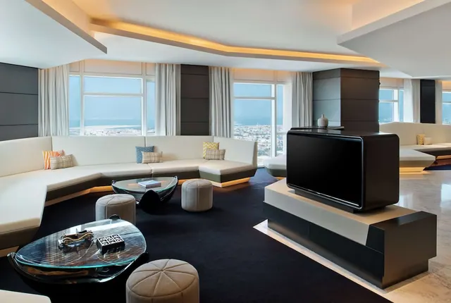 A living area with a curved white sofa, glass coffee tables, a large TV on a black cabinet, and windows looking over the city and sea.
