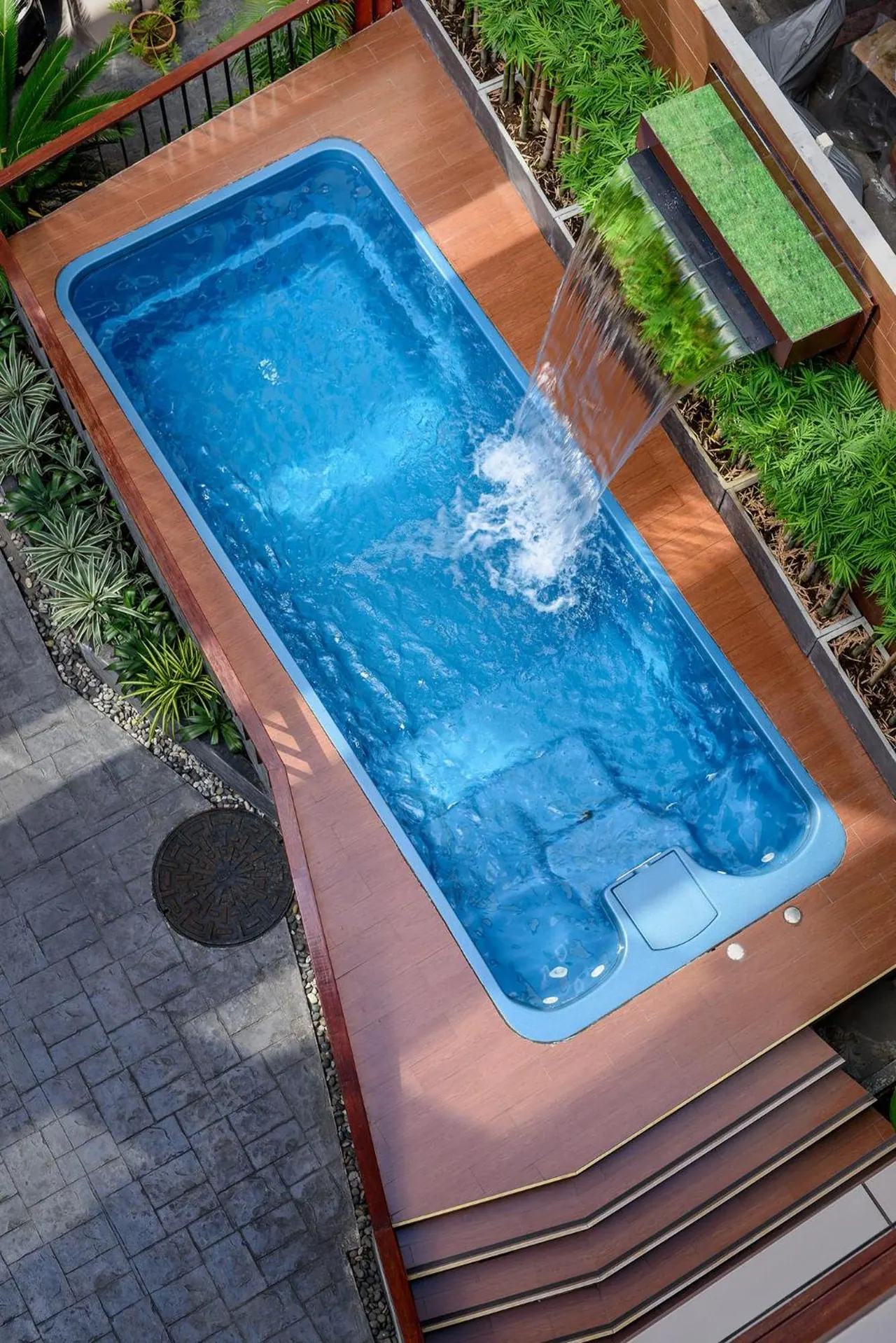 Overhead view of a small rectangular pool with a waterfall spout on a wooden deck, bordered by plants and stone paving.