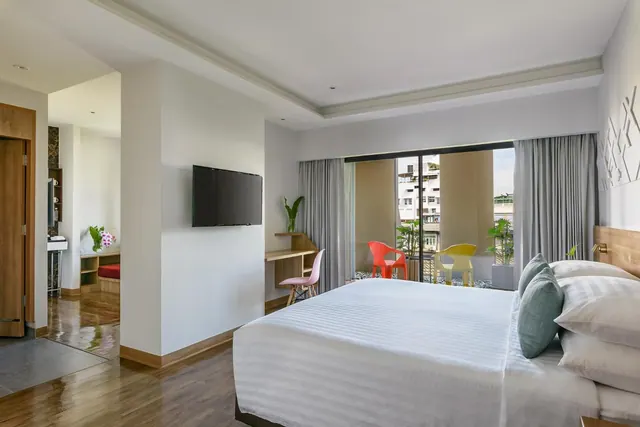 A large white bed sits in a bedroom beside a wall-mounted television, a desk with a pink chair, and a sliding glass door onto a balcony with coloured chairs and nearby buildings beyond.
