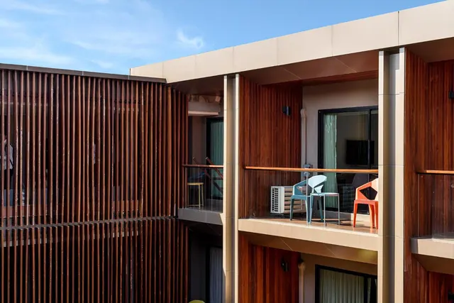 Exterior of a multi-storey building with wooden slatted walls, a balcony holding blue and orange plastic chairs, and a wall-mounted air conditioner unit under a clear blue sky.