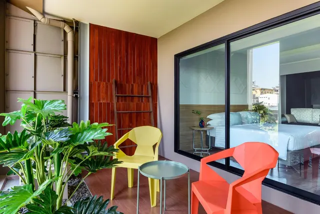 A balcony with a yellow and an orange plastic chair, a round grey side table, potted plants, and a large glass door showing a bedroom with a made bed.