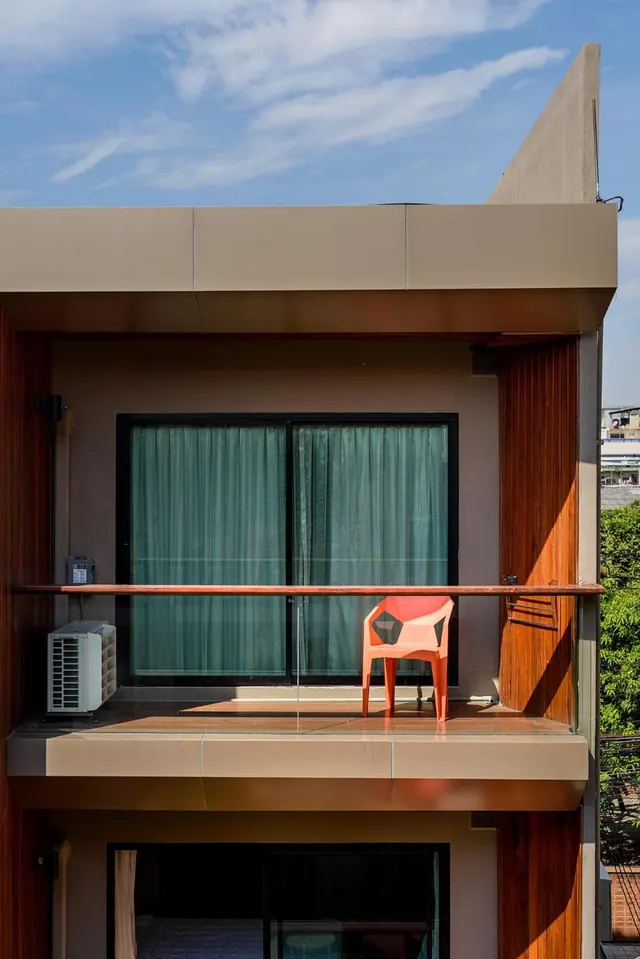 A balcony with a glass railing and an orange plastic chair sits beside a sliding door with teal curtains, with a wall-mounted air conditioner unit on the left.