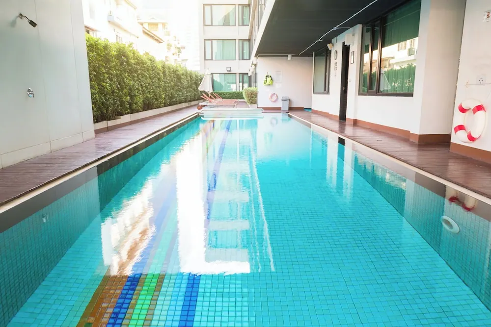 An outdoor lap pool with blue mosaic tiles runs beside a wooden deck, with sun loungers and a parasol at the far end, a hedge and building walls behind.