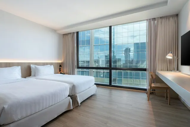 A twin-bedded hotel room with floor-to-ceiling windows looking out onto high-rise glass buildings, with a desk, desk lamp and wall-mounted television on the right.