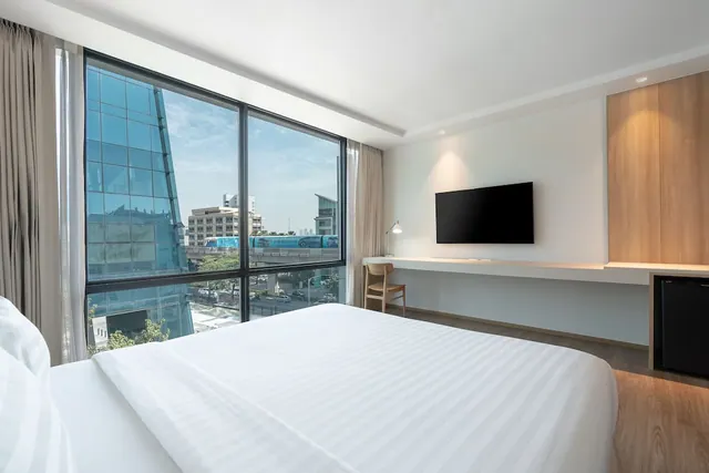 A bedroom with a made king bed facing a large window that overlooks city buildings and an elevated train line, with a wall-mounted television, a desk with chair, and a minibar.