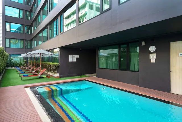 A rectangular pool with rainbow-striped tiles on its entry steps sits beside a row of sun loungers and striped umbrellas on artificial turf.