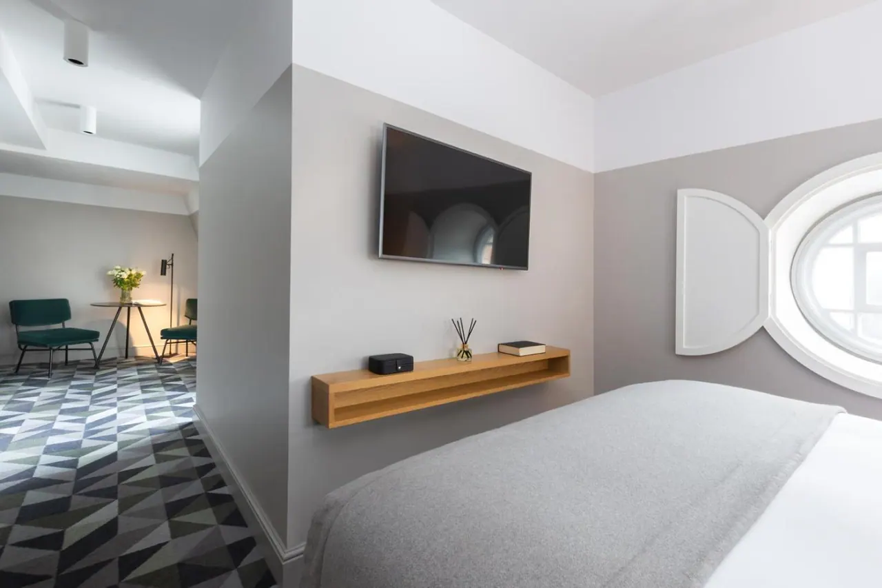A bedroom with a made bed, a wall-mounted television, a wooden floating shelf holding a diffuser and a book, and a seating area with a green armchair beyond.