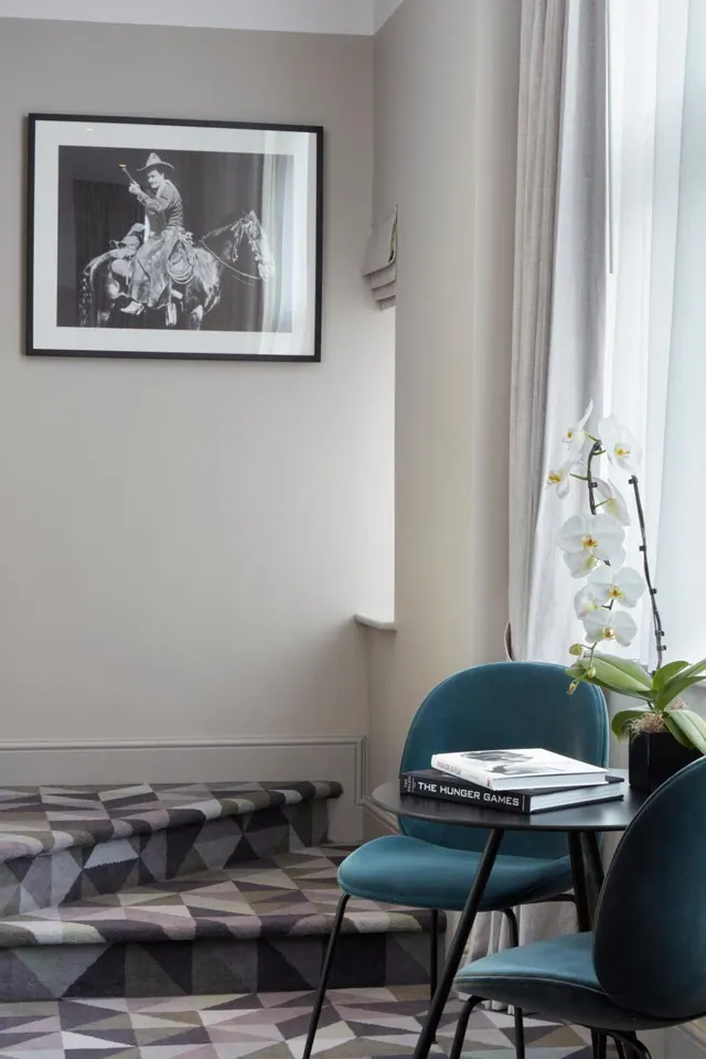 A room corner shows a framed black and white photograph on a pale wall, a geometric patterned carpeted step, two teal chairs at a round table holding books and a white orchid, and a window with sheer curtains.