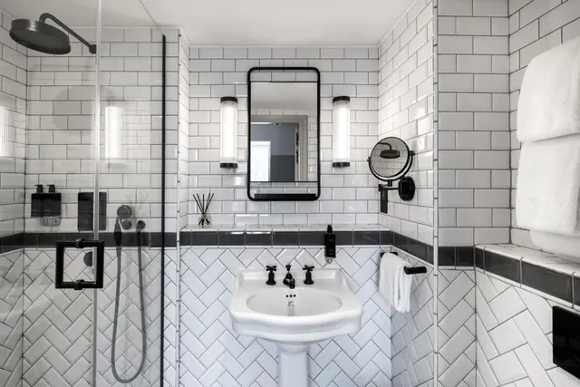 A white subway-tiled bathroom with a glass-enclosed walk-in shower, a pedestal sink with black taps, a framed wall mirror, and folded towels on a rail.