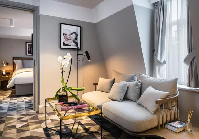 A living area with a grey sofa and cushions, a glass coffee table holding an orchid and a book, and a bed visible through the open doorway on the left.
