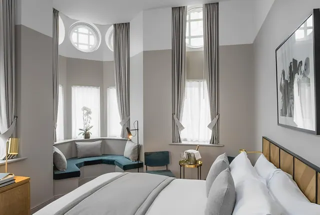 A bedroom with a large bed with white linen, a teal window seat beneath tall curtained windows, and a framed black and white photograph on the wall.