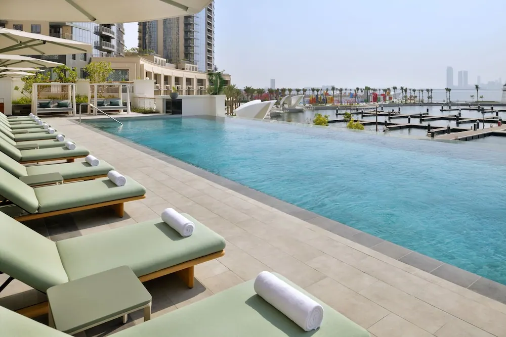 An infinity-edge pool with green sun loungers and rolled white towels overlooks a marina with docks and high-rise towers in the distance.