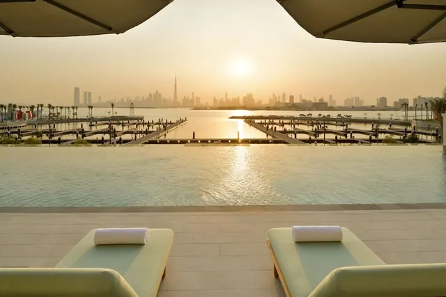 An infinity-edge pool with two sun loungers and rolled white towels overlooks a marina and the Dubai skyline, including the Burj Khalifa, at sunset.