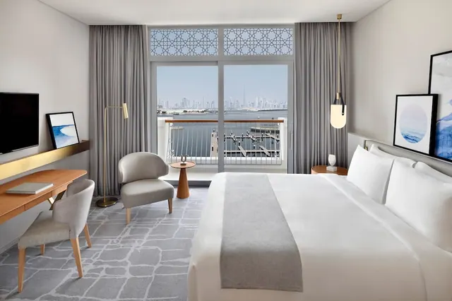 A bedroom with a large white bed, a wooden desk and an armchair, with a glass door opening onto a balcony that overlooks water toward a city skyline.