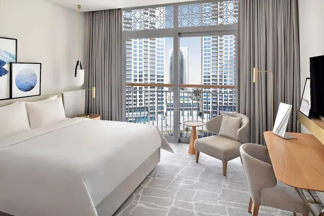 A hotel bedroom with a king bed, two armchairs, a wooden desk and a television, with a glass door opening onto a balcony overlooking high-rise towers.