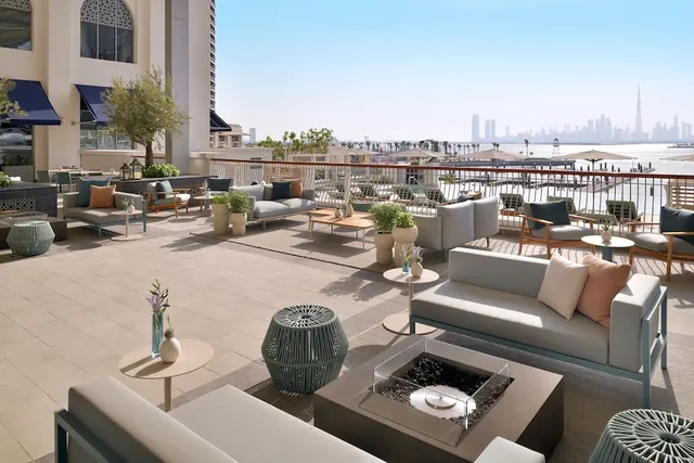 An outdoor terrace with lounge sofas and a fire pit overlooks calm water and the Dubai skyline, with the Burj Khalifa visible in the distance.