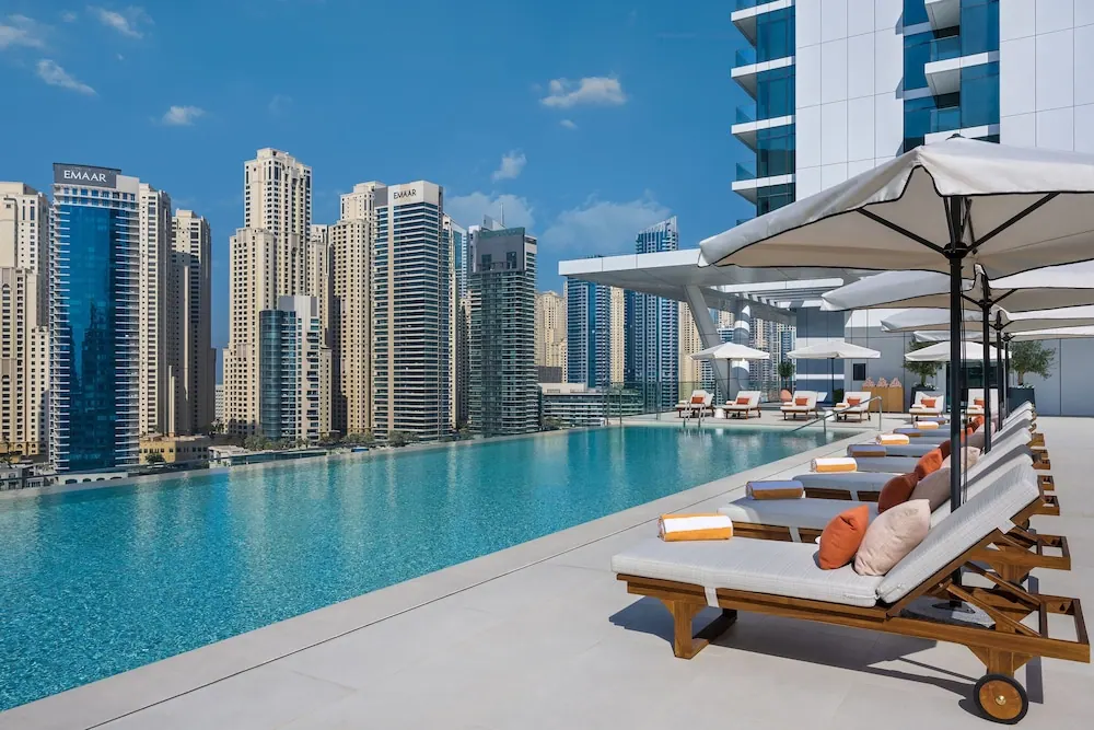 A rooftop swimming pool with white shade umbrellas and cushioned sun loungers faces a skyline of high-rise towers under a blue sky.