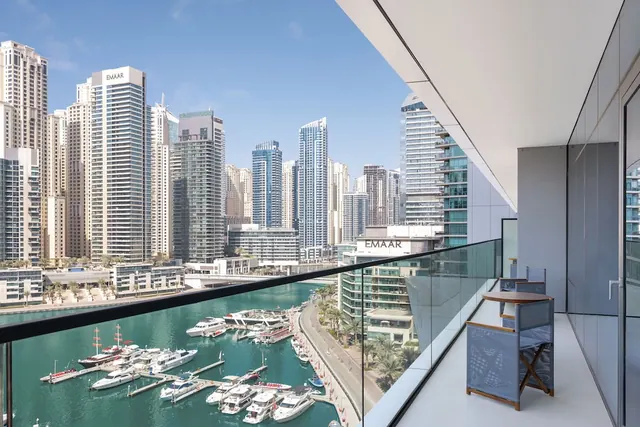 A balcony with a glass railing, two folding chairs and a small table overlooks a marina with moored yachts and high-rise towers.